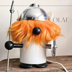 Könnte beinhalten: Eine weiße Keramiklampe in Form eines Wikingers mit einem silbernen Helm, orangefarbenem Fell und einem schwarzen Auge. Die Lampe hat einen schwarzen Griff und einen silbernen Schild. Die Lampe ist mit "OLAF" beschriftet.