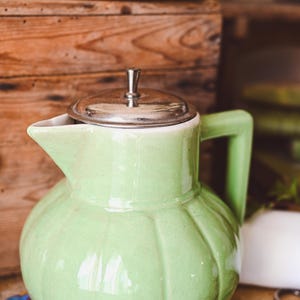 Op de afbeelding: Een groene keramische kan met een zilveren deksel en een handvat. De kan heeft een rond lichaam met een getextureerd oppervlak.