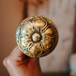 Op de afbeelding: Een ronde koperen deurknop met een verhoogd, in reliëf gemaakt bloemmotief met vijf afzonderlijke bloemblaadjes die vanuit een centraal punt uitstralen. Het metalen oppervlak heeft een gepolijste afwerking met donkere, verdiepte delen.