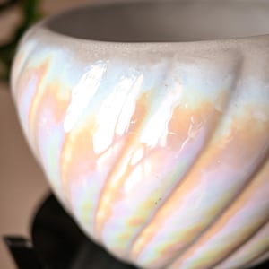 Può includere: Una ciotola in ceramica rotonda con una finitura lucida e iridescente e creste diagonali a spirale. La superficie riflette la luce in tenui tonalità pastello di rosa, blu e oro, creando un effetto scintillante sull'esterno curvo e scanalato.