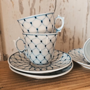 Può includere: Tazze e piattini bianchi impilati con un motivo a reticolo blu. Le tazze hanno manici decorati e sono posizionate su una superficie di legno. Il design si ripete sui piattini, creando un set coordinato.