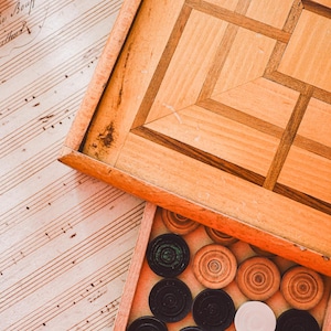 Op de afbeelding: Een houten backgammonbord met een geometrisch ontwerp, gedeeltelijk open om een lade met spelstukken te onthullen. De stukken zijn zwart, bruin en wit. Het bord rust op een vel oud bladmuziek.