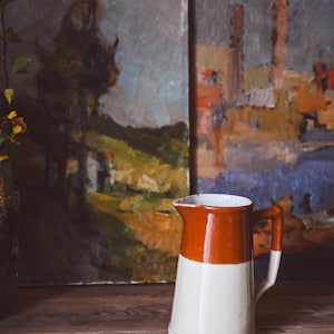 Op de afbeelding: Een witte keramische kan met een bruine bovenkant, die op een houten oppervlak staat voor twee schilderijen.