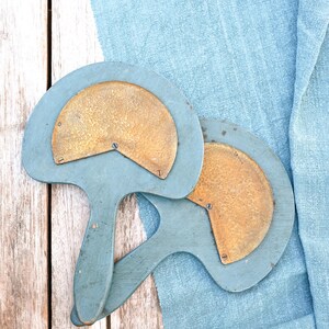 Puede incluir: Dos herramientas de mano en forma de abanico con mangos y marcos de madera pintados de azul. Cada herramienta presenta una placa de latón martillado en forma de semicírculo fijada al centro con pequeños tornillos metálicos. Las herramientas están superpuestas.