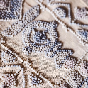 May include: Close-up of a beige fabric with detailed beadwork. The beads are in blue, white, and pale pink, forming floral and geometric designs. The fabric's texture is evident, creating a delicate, vintage aesthetic.