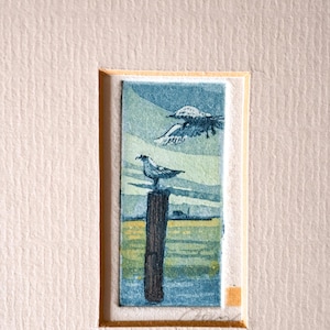 Puede incluir: Una pequeña impresión rectangular que muestra un poste de madera marrón oscuro en un mar azul y amarillo. Un ave se posa sobre el poste mientras otra vuela arriba contra un cielo pálido. La obra está montada sobre papel texturizado color crema.