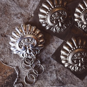 Puede incluir: Un adorno de metal plateado con un diseño decorativo y un juego de llaveros plateados. El adorno está sobre una superficie marrón texturizada.