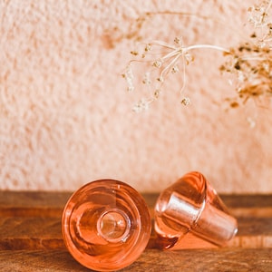 May include: Two clear, pink glass bottle stoppers rest on a wooden surface. One stopper is upright, while the other is on its side. The background features a textured, peach-colored wall and a sprig of dried flowers.