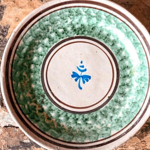 Puede incluir: Un plato de cerámica con un diseño pintado a mano. El plato presenta un borde verde moteado, un borde marrón y un círculo central blanco con un motivo floral azul. El plato descansa sobre una superficie de madera rústica.
