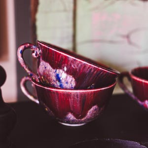 Peut inclure: Deux tasses à thé en céramique bordeaux empilées, avec des anses. Les tasses ont une finition brillante avec des mouchetures bleues et blanches. Les tasses sont sur une surface sombre, avec une troisième tasse visible en arrière-plan.
