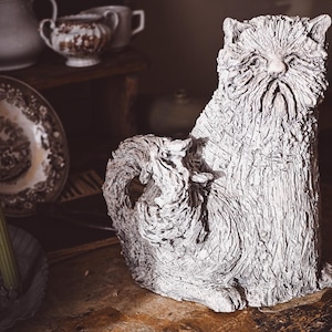 Puede incluir: Escultura de gato texturizada, blanquecina, con superficie rugosa y detallada. El gato está sentado, con rostro prominente y cola enroscada. El fondo incluye vajilla antigua y un candelabro.