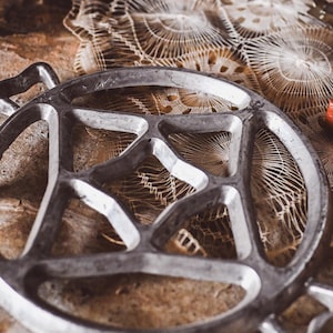 Könnte beinhalten: Ein silbernes Metallwerkzeug zum Herstellen von Spitze mit einer runden Form und einem Sternmuster in der Mitte. Das Werkzeug liegt auf einer braunen Oberfläche mit einer weißen Spitzendeckchen im Hintergrund.