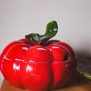 Pode incluir: Um prato de servir de cerâmica vermelha em forma de tomate com uma tampa de folha verde. O prato está sobre uma mesa de madeira.