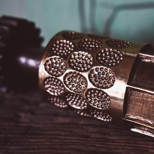 May include: Close-up of a brass metal embossing roller with a repeating pattern of small, raised circles. The roller is on a wooden surface, with a blurred teal background. The tool is used for adding texture to paper or other materials.