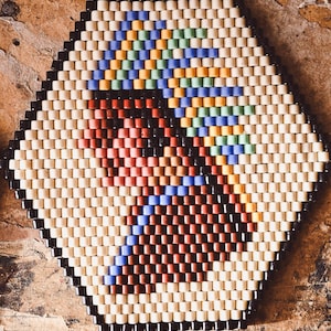 May include: A beaded coaster with a geometric design. The coaster features a black border and a central pattern of cream, red, blue, green, and yellow beads. The coaster is approximately 15 cm in diameter.