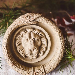 Peut inclure: Objet décoratif circulaire beige avec une tête de lion au centre, entourée d'un serpent se mordant la queue. Le serpent et le lion sont en relief, avec des textures détaillées. L'objet est probablement en pierre ou dans un matériau similaire.