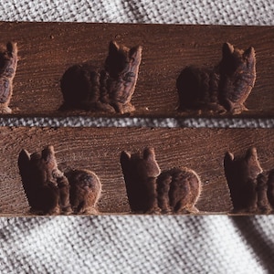 May include: Two wooden decorative strips with carved brown cat figures. The cats are in a sitting position, with their tails curled. The wood has a rustic, textured appearance. The background is a white, textured fabric.