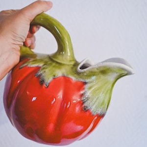 Puede incluir: Jarra de cerámica con forma de tomate, con cuerpo rojo brillante y detalles verdes. El asa y la parte superior son verdes, el pico es blanco. La jarra está sujeta por una mano. Capacidad aproximada de 1 litro.