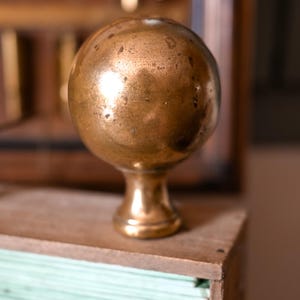 May include: A spherical brass knob with a polished, metallic finish and a tapered base. The surface of the sphere shows subtle signs of wear and natural patina, featuring a smooth, rounded top and a sturdy, narrow neck that connects to a circular pedestal.