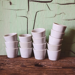 Puede incluir: Un conjunto de tres pilas de tazas de cerámica blanca, cada una con un labio ligeramente redondeado, dispuestas sobre una superficie de madera. Las tazas están apiladas en un patrón escalonado, creando una exhibición visualmente atractiva.