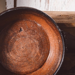 May include: A rustic, round, earthenware bowl with a dark brown rim and a reddish-brown interior. The bowl has a textured surface with visible wear and tear, suggesting age and use. The bowl is likely used for serving or display.