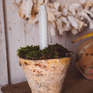 Op de afbeelding: Een rustieke, verweerde terracotta pot gevuld met groen mos, met een lange, witte, spiraalvormige kaars. De pot staat op een houten oppervlak, met een vage achtergrond met een decoratieve krans.