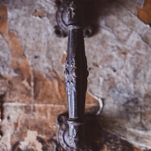 Peut inclure: Une poignée de porte en métal noir avec des détails ornés. La poignée est fixée à un mur avec de la peinture écaillée.