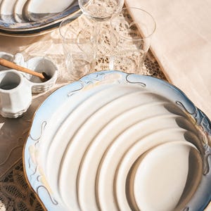 Könnte beinhalten: Eine Sammlung eleganter Tischutensilien, darunter eine große weiße und blaue Servierplatte mit gewelltem Rand, mehrere klare Trinkgläser und kleine weiße Servierschalen. Die Teller haben einen goldenen Rand.