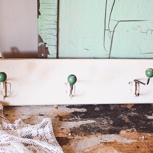 Puede incluir: Un perchero de madera blanco vintage con tres ganchos de metal y pomos verdes en forma de bola. El perchero está montado sobre una superficie de madera rústica, con un paño de encaje en primer plano y una tabla verde desgastada.