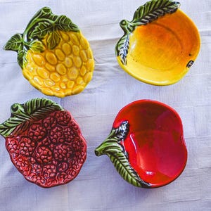 Puede incluir: Cuatro platos de cerámica con forma de fruta: una piña amarilla, un mango amarillo, una frambuesa roja y una manzana roja. Cada plato tiene un acento de hoja verde. Los platos están dispuestos sobre una superficie blanca texturizada.