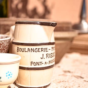 May include: Cream-colored ceramic pitcher with a dark brown rim and bands, featuring the text "BOULANGERIE - PAT J. RISSE PONT-A-MOUSSON" in black. A small, decorated cup is in the foreground. The pitcher has a barrel-like shape.