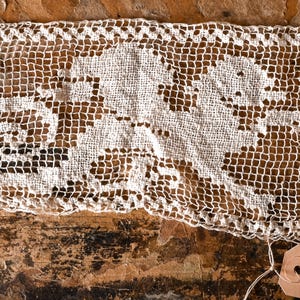 May include: A rectangular strip of off-white lace featuring a repeating woven pattern of a stylized creature with a curled tail. The lace has a grid-like structure and is attached to a small tan paper tag with the text zap printed in black ink.