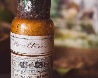 May include: A vintage glass bottle with a rusty metal cap and a green label. The label reads "Me altose" and "Droguerie" with the name "DARRASSE FRERES" in a larger font. The bottle is sitting on a dark surface.