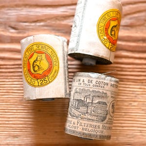 Könnte beinhalten: Drei zylindrische Spulen aus belgischem Leinenfaden, umwickelt mit cremefarbenen Papieretiketten. Zwei Etiketten zeigen ein gelbes kreisförmiges Emblem mit einer schwarzen Katzenillustration und dem Text THREAD FOR SEWING MACHINE.