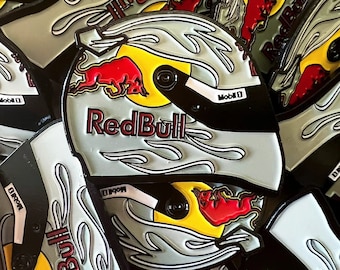 Könnte beinhalten: Eine Sammlung von Red Bull Rennhelm-förmigen Pins. Die Pins zeigen ein graues Helmdesign mit Flammenakzenten, dem Red Bull Logo und dem Mobil 1 Logo. Das Visier des Helms ist schwarz, und der Hintergrund ist unscharf.