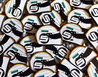 May include: A close-up of a pile of white and black enamel pins shaped like racing helmets. Each helmet has a number "5" on the side and a green stripe across the top.