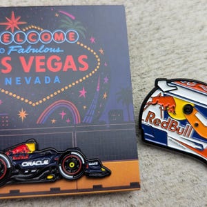 Könnte beinhalten: Ein Pin-Set mit einem Las Vegas-Themen-Design und einem Red Bull-Rennhelm. Der Las Vegas-Pin hat den Text "Welcome to Fabulous Las Vegas Nevada" und ein Rennwagen. Der Helm-Pin hat das Red Bull-Logo.