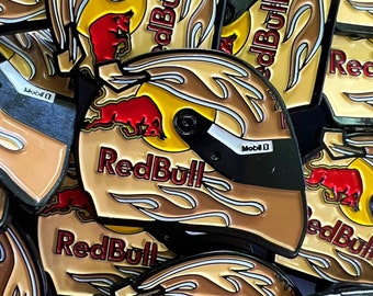 Könnte beinhalten: Eine Sammlung von Red Bull Rennhelm-Anstecknadeln. Die Anstecknadeln haben ein braunes, gelbes und rotes Design mit dem Red Bull-Logo und Flammendetails. Die Anstecknadeln tragen die Aufschrift "Red Bull" und "Mobil 1".