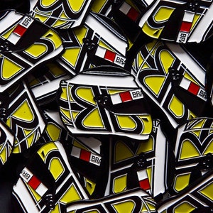 Op de afbeelding: Een close-up van een stapel zwarte en gele emaillepins met een wit en rood ontwerp. De pins tonen een gestileerde helm met het woord "BRA" erop.