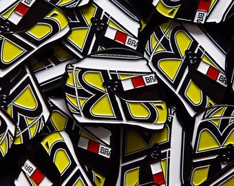 May include: A close-up of a pile of black and yellow enamel pins with a white and red design. The pins feature a stylized helmet with the word "BRA" on it.