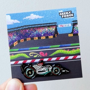 May include: A silver and black Formula 1 race car pin with the text "Mexico" on the background. The pin is from The Geeky Forge.