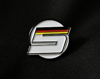 May include: A white enamel pin with a black outline, featuring the number "5" in a stylized font. The number is layered with the colors of the German flag: black, red, and yellow.