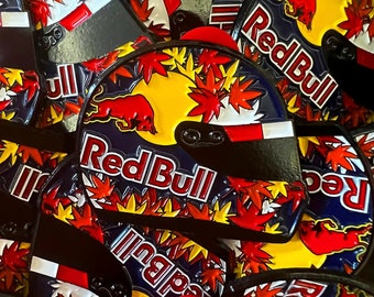 Könnte beinhalten: Eine Sammlung von Red Bull Rennhelm-förmigen Pins. Die Pins zeigen das Red Bull Logo, einen Bullen und die Worte "Red Bull" in Rot. Die Helme sind mit roten und gelben Ahornblättern vor einem gelben und blauen Hintergrund verziert.