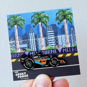 May include: A black and orange Formula 1 race car pin with a pixelated city skyline and palm trees in the background. The pin says "Melbourne" on the bottom.