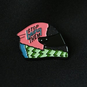 Può includere: Uno spillo in smalto nero e rosa a forma di casco da corsa con il testo "STOP being THEM" sul lato.