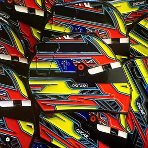 Puede incluir: Primer plano de una pila de pines de colores con forma de casco con la palabra "OSCAR" impresa en cada uno. Los pines presentan un esquema de colores negro, rojo, amarillo, azul y blanco con un diseño de bandera a cuadros.