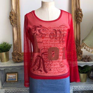 May include: Red sheer long-sleeved shirt with a graphic print of the word "LOVE" in a stylized font with a gold background. The shirt has a round neckline.
