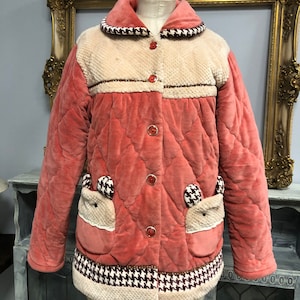May include: A quilted pink jacket with a white collar and trim. The jacket has a button closure and two pockets with bear ears.