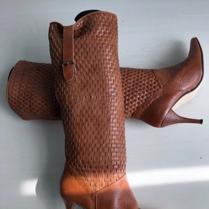 以下が含まれることがあります： 編み込み模様の入った茶色の革のニーハイブーツ。ポインテッドトゥで、高いヒールとサイドにバックルが付いています。