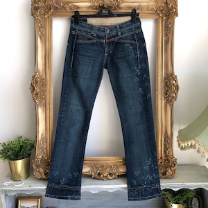 May include: Dark wash denim jeans with floral embroidery and a distinctive waist design. The jeans have a straight leg cut and are displayed on a hanger. The jeans have a red accent on the waist.